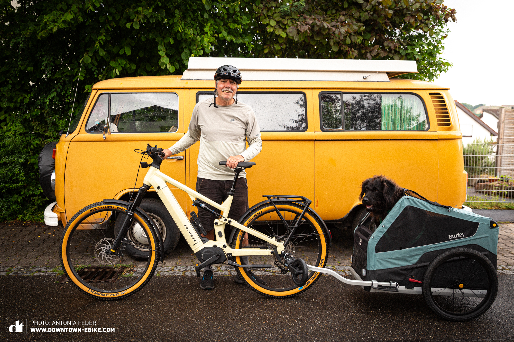 Manne steht mit Fahrrad und Fahrrad-Hundeanhänger vor einem VW Bus und hat dieselbe Länge. 