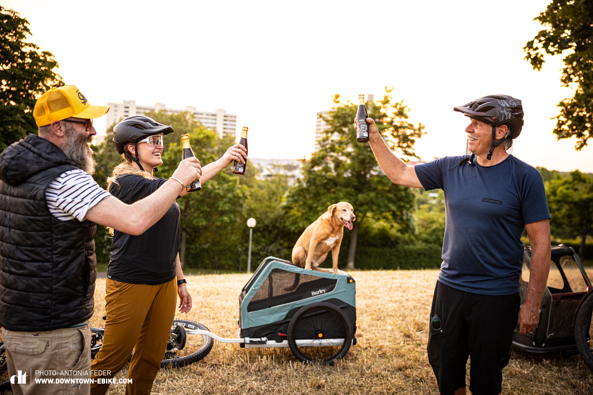 Hier sieht man wie Oli, Antonia und Manne mit einem Bier in der Luft anstoßen. Sie stehen auf einem Feld und im Hintergrund sitzt Leo auf dem Dach eines Fahrrad-Hundeanhänger. 