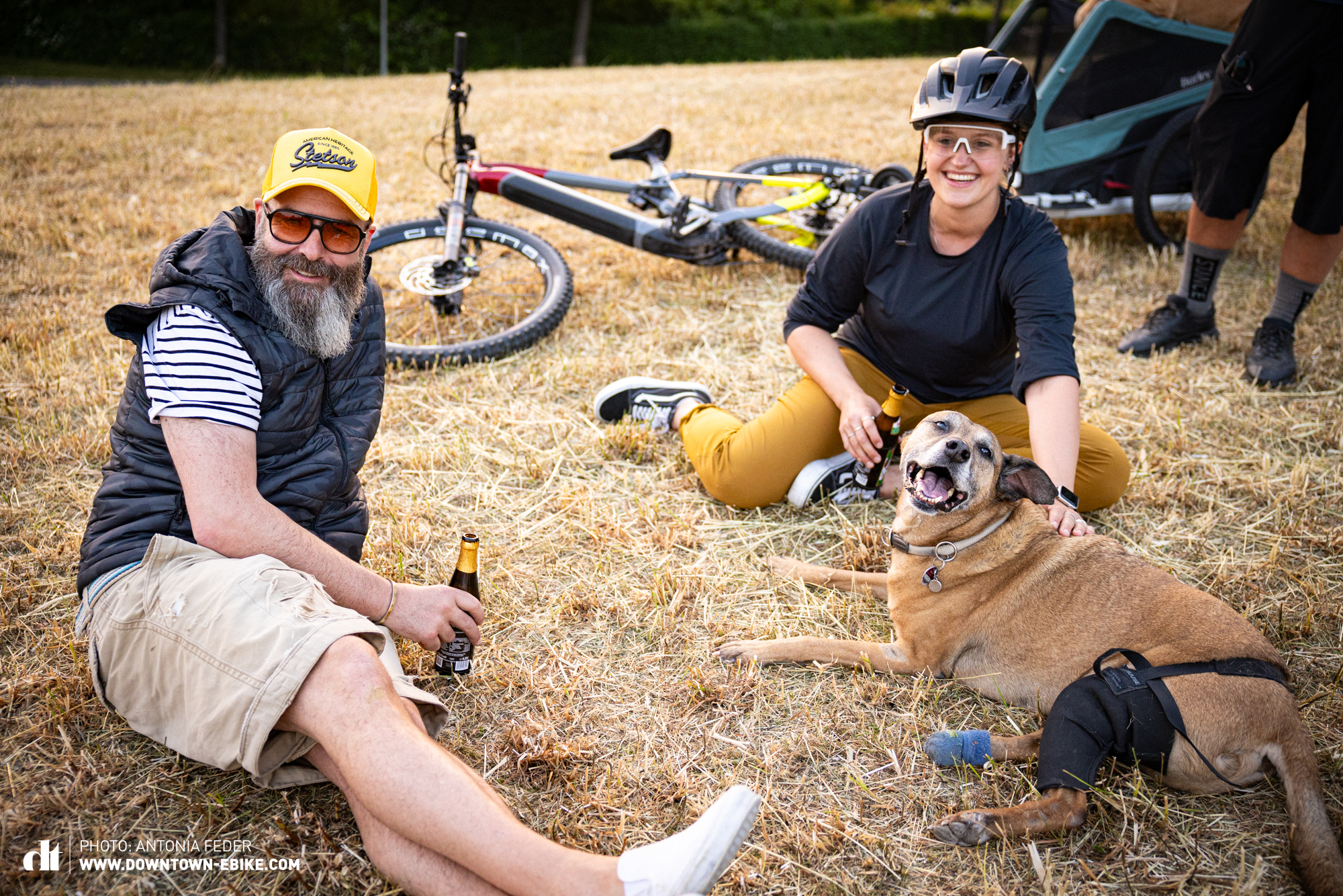Nachdem die Tour mit dem Fahrrad-Hundeanhänger geschafft ist, sitzen Oli und Antonia mit Bruce auf dem Feld und genießen ihr Bier. Bruce schaut happy in die Kamera. 